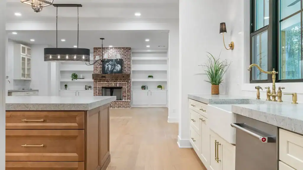 293741fffa7e92c7db2c90b1a77d8c4e-uncropped_scaled_within_1536_1152 Bright modern kitchen with white cabinetry, gold hardware, granite countertops, and a farmhouse sink. Spacious open layout with brick fireplace in the background.