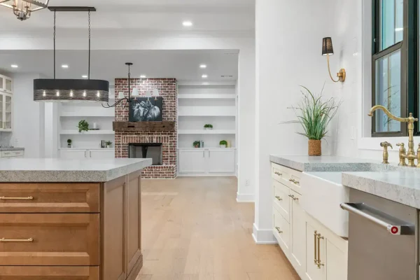 Bright modern kitchen with white cabinetry, gold hardware, granite countertops, and a farmhouse sink. Spacious open layout with brick fireplace in the background.