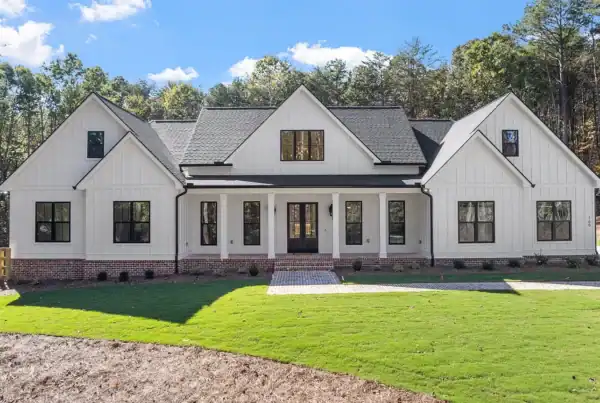 Modern white farmhouse with black accents and expansive front porch, surrounded by lush greenery and new landscaping.
